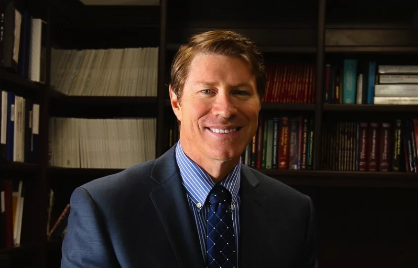 A man in a suit and tie smiles in front of a bookcase decorated with medical books, suggesting expertise in breast augmentation surgery.
