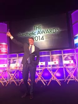 A Las Vegas surgeon in a suit standing on stage with a trophy.
