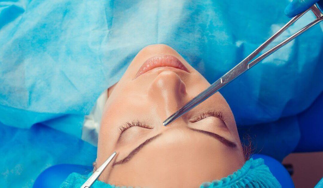 Close-up of a person lying on an operating table, preparing for rhinoplasty, wearing a blue surgical gown and cap, with surgical tools positioned near the face.