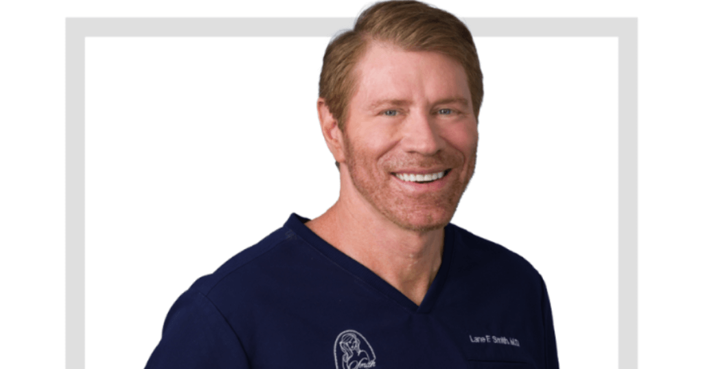 Male Gynecomastia Surgery in Las Vegas: What Men Need to Know 1 A man with light brown hair and a beard, smiling, wearing navy blue medical scrubs with embroidered logos, stands in front of a plain background—representing Male Gynecomastia Surgery for men in Las Vegas.