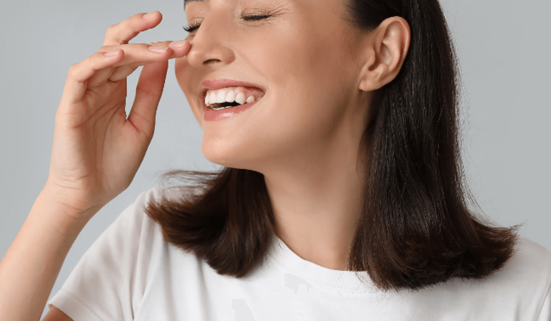 Woman with shoulder-length brown hair smiling and pinching her nose, wearing a white t-shirt against a neutral background—perfect for discussing rhinoplasty cost in Las Vegas.