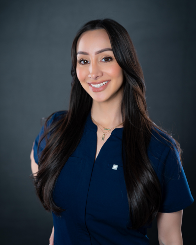 Woman with long dark hair wearing a navy blue scrub top, smiling and standing against a plain dark background. Woman with long dark hair wearing a navy blue scrub top, smiling and standing against a plain dark background.