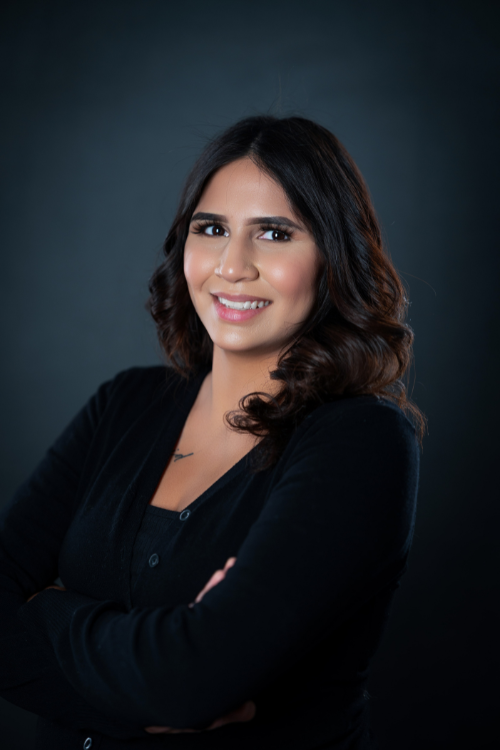 A woman with medium-length dark hair smiles at the camera, wearing a black top and standing against a plain dark background with her arms crossed. A woman with medium-length dark hair smiles at the camera, wearing a black top and standing against a plain dark background with her arms crossed.