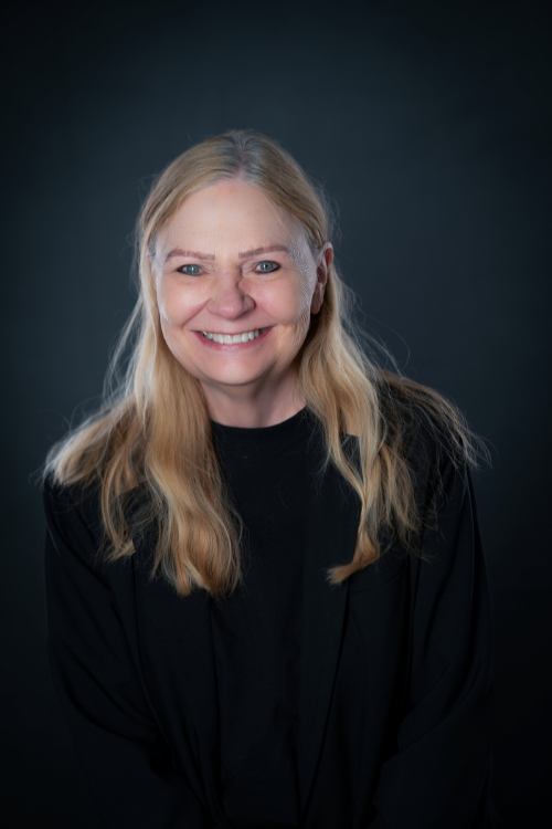 A middle-aged woman with long blonde hair and a black top smiles in front of a dark background. A middle-aged woman with long blonde hair and a black top smiles in front of a dark background.
