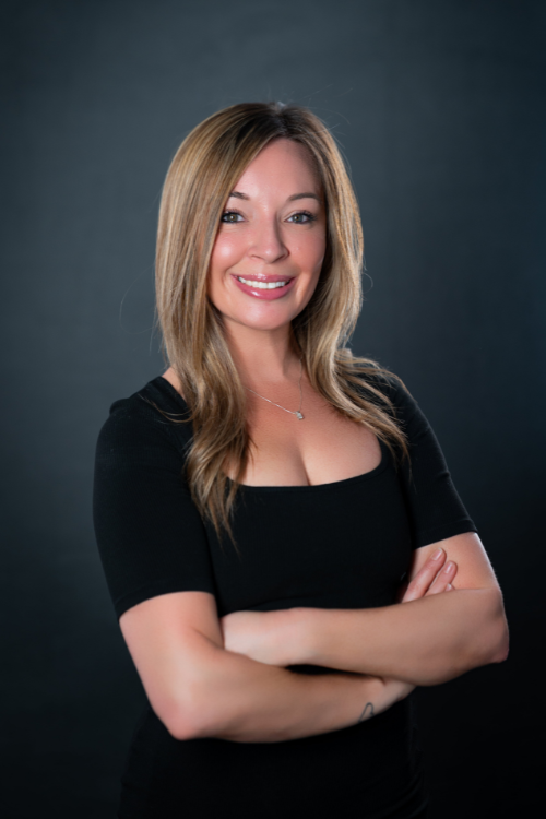 Woman with long light brown hair wearing a black short-sleeve top stands with arms crossed, smiling in front of a dark background. Woman with long light brown hair wearing a black short-sleeve top stands with arms crossed, smiling in front of a dark background.