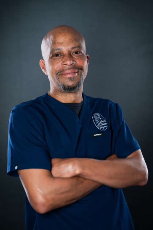 A man wearing navy blue medical scrubs stands against a dark background with his arms crossed and a slight smile. A man wearing navy blue medical scrubs stands against a dark background with his arms crossed and a slight smile.
