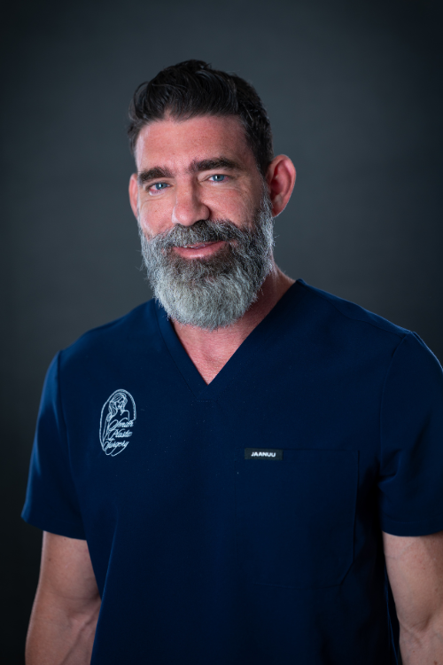 A man with a gray beard and short hair wearing navy blue medical scrubs stands facing the camera against a dark background. A man with a gray beard and short hair wearing navy blue medical scrubs stands facing the camera against a dark background.