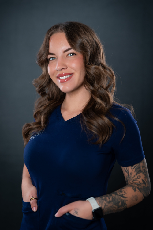 A woman with long wavy brown hair, wearing a navy blue top and a smartwatch, stands against a dark background, smiling with her hands in her pockets. A woman with long wavy brown hair, wearing a navy blue top and a smartwatch, stands against a dark background, smiling with her hands in her pockets.