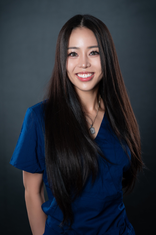 A woman with long dark hair wearing blue scrubs smiles at the camera against a plain dark background. A woman with long dark hair wearing blue scrubs smiles at the camera against a plain dark background.