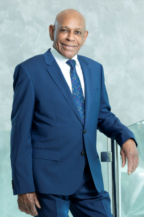 An older man in a blue suit and patterned tie stands indoors, smiling, with one hand resting on a glass railing. An older man in a blue suit and patterned tie stands indoors, smiling, with one hand resting on a glass railing.