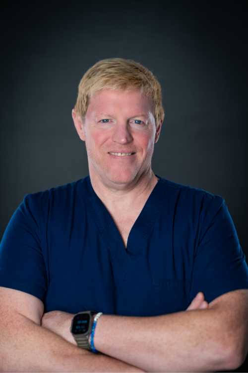 A man with light hair wearing navy blue scrubs and a smartwatch stands with arms crossed, smiling against a dark background. A man with light hair wearing navy blue scrubs and a smartwatch stands with arms crossed, smiling against a dark background.