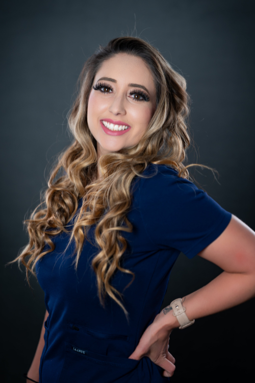 Woman with long, wavy blonde hair wearing a navy blue top and a beige watch, smiling and posing with one hand on her hip against a dark background. Woman with long, wavy blonde hair wearing a navy blue top and a beige watch, smiling and posing with one hand on her hip against a dark background.