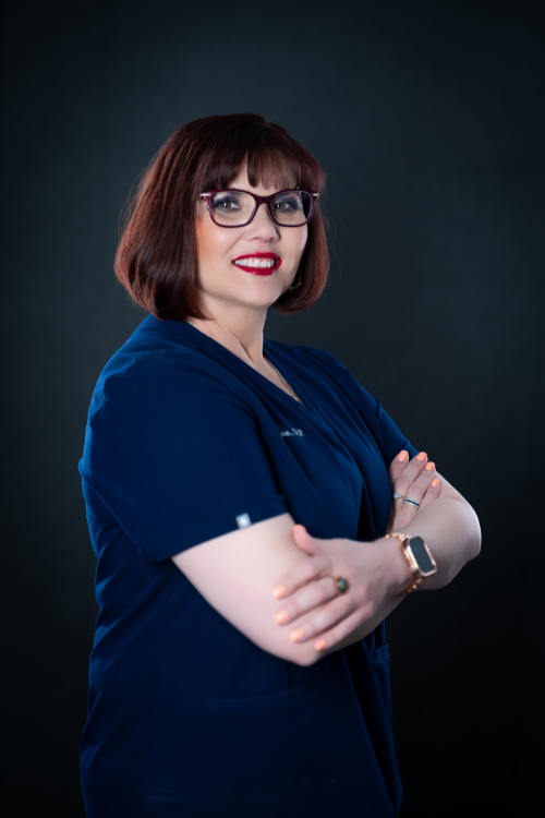 A woman with short brown hair wearing glasses and blue scrubs stands with arms crossed, smiling against a dark background. A woman with short brown hair wearing glasses and blue scrubs stands with arms crossed, smiling against a dark background.