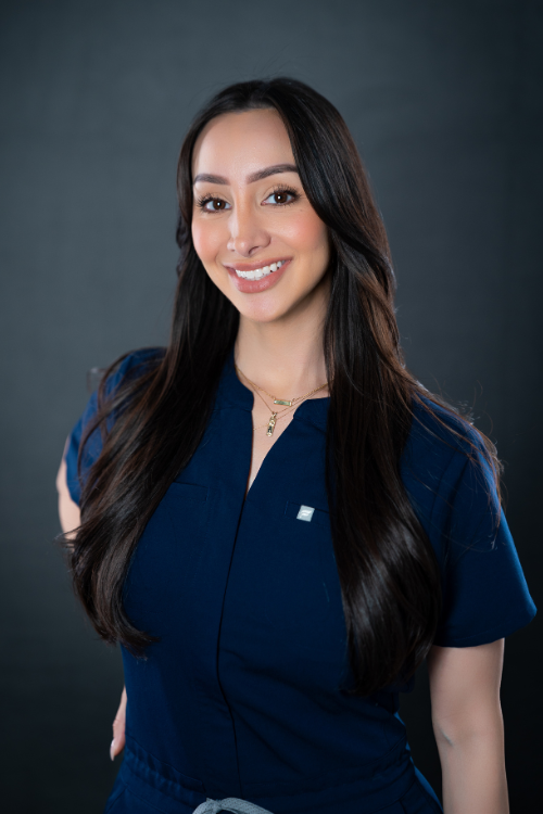 Woman with long dark hair wearing a navy blue scrub top stands against a dark background, smiling at the camera. Woman with long dark hair wearing a navy blue scrub top stands against a dark background, smiling at the camera.