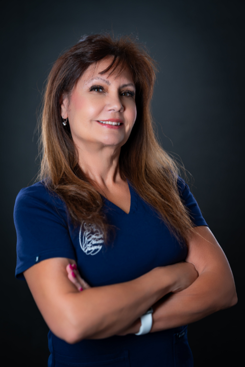 Woman with long brown hair wearing a navy blue shirt stands with arms crossed, smiling, against a dark background. Woman with long brown hair wearing a navy blue shirt stands with arms crossed, smiling, against a dark background.