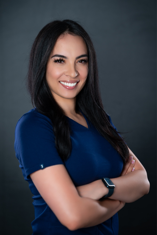 Woman with long dark hair wearing a navy blue top and smartwatch stands against a dark background, smiling with arms crossed. Woman with long dark hair wearing a navy blue top and smartwatch stands against a dark background, smiling with arms crossed.