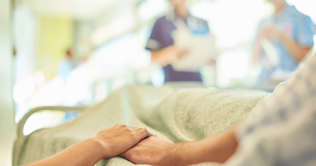How to Minimize Scars and Support Healing After Cosmetic Surgery 1 A person lies in a hospital bed, holding hands with a visitor. Medical staff stand in the blurred background, suggesting a healing environment after cosmetic surgery.