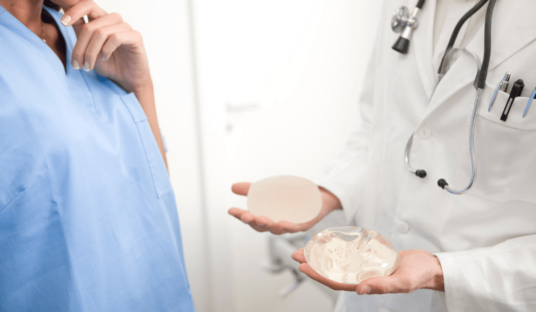 A doctor in a white coat shows two types of breast implants to a person in blue scrubs during a breast augmentation consultation in Las Vegas.