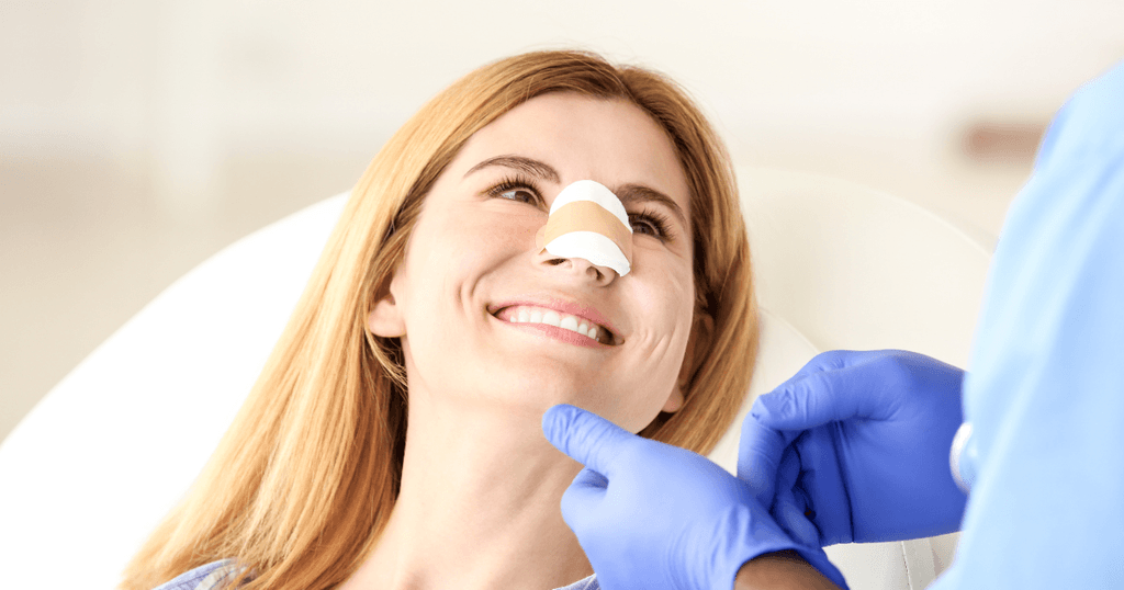 How Much Is a Nose Job in Las Vegas? Factors That Influence Rhinoplasty Cost 1 Smiling woman with bandages on her nose sits as a person in blue gloves examines her chin in a medical setting, possibly discussing rhinoplasty cost after undergoing a nose job Las Vegas patients often seek.